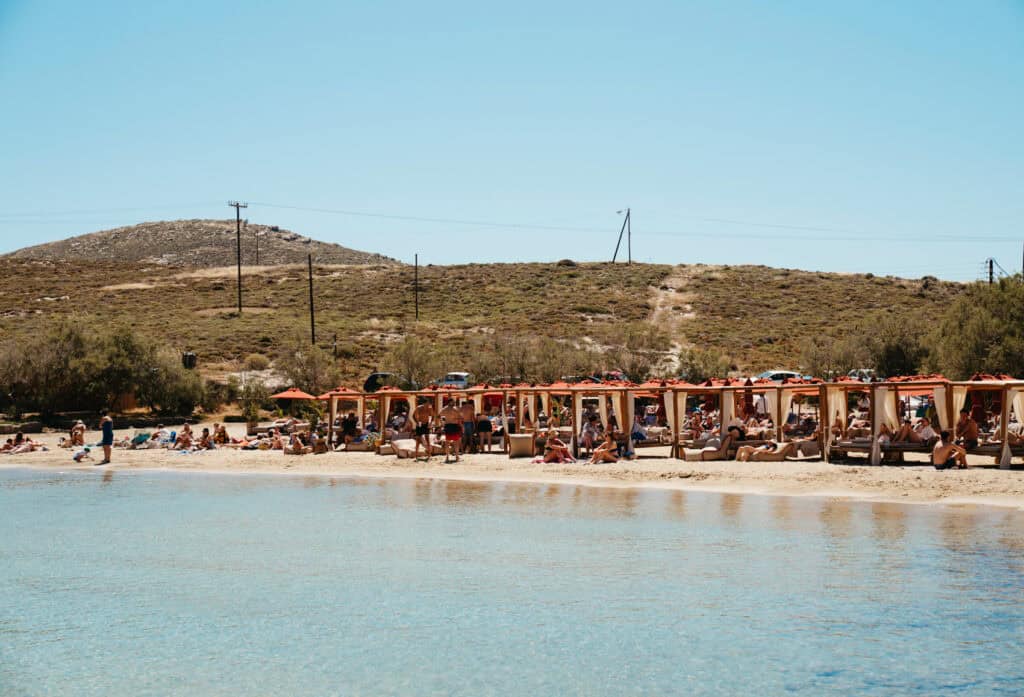 View of Monastiri Beach, Paros, from the water, showing rows of shaded cabanas and sunbeds filled with beachgoers, set against a dry hillside landscape.