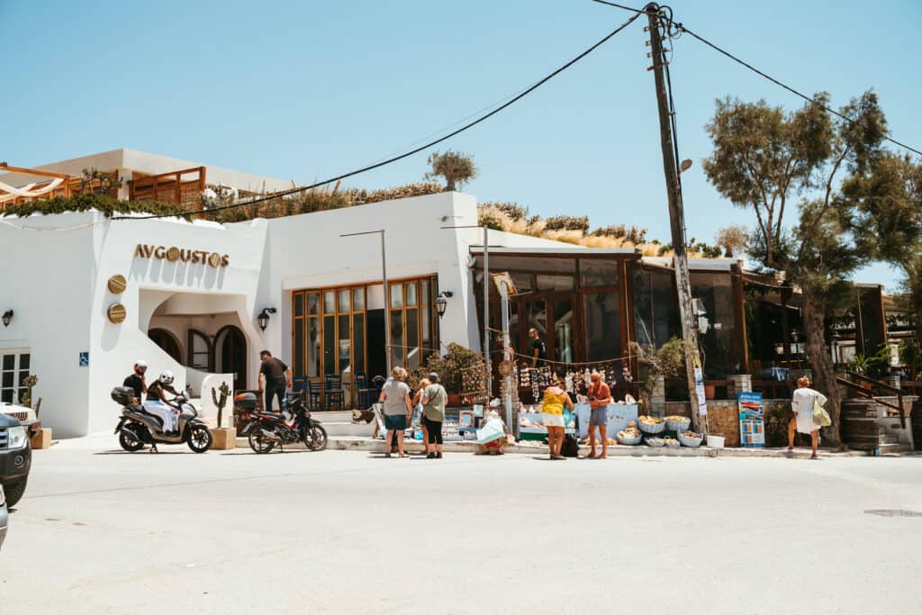 Street view of Avgoustos restaurant in Agia Anna, Naxos, with people browsing local souvenirs and riding scooters under a clear blue sky.