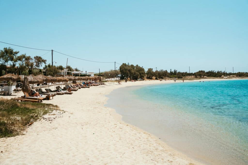 Quiet stretch of Agia Anna Beach in Naxos, lined with thatched umbrellas and wooden loungers along soft golden sand and shallow turquoise waters.