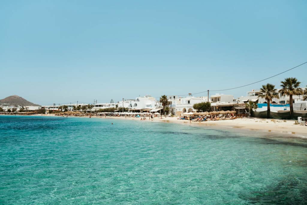 Panoramic view of Agia Anna Beach in Naxos, lined with whitewashed buildings, beach bars, and sunbeds along the clear turquoise shoreline.
