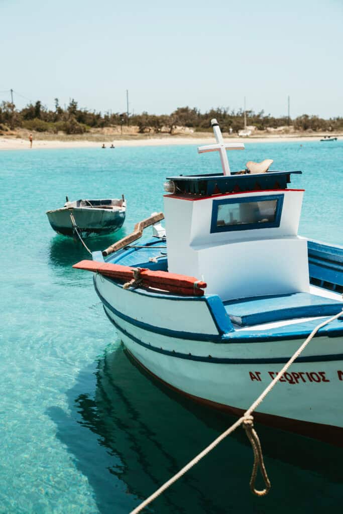 Traditional white and blue fishing boat tied in the shallow turquoise waters of Agia Anna, Naxos, with a quiet beach and trees in the background.