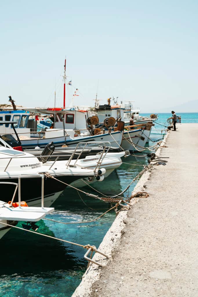 Fishing boats tied along the dock in Agia Anna, Naxos, with a man working on the pier beside clear turquoise water and a bright summer sky.