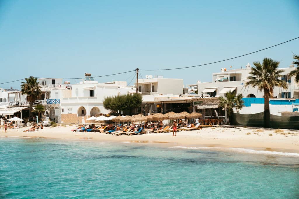 Agia Anna Beach in Naxos with sunbeds and straw umbrellas lining the sandy shore, backed by white Cycladic buildings, palm trees, and beachfront tavernas.