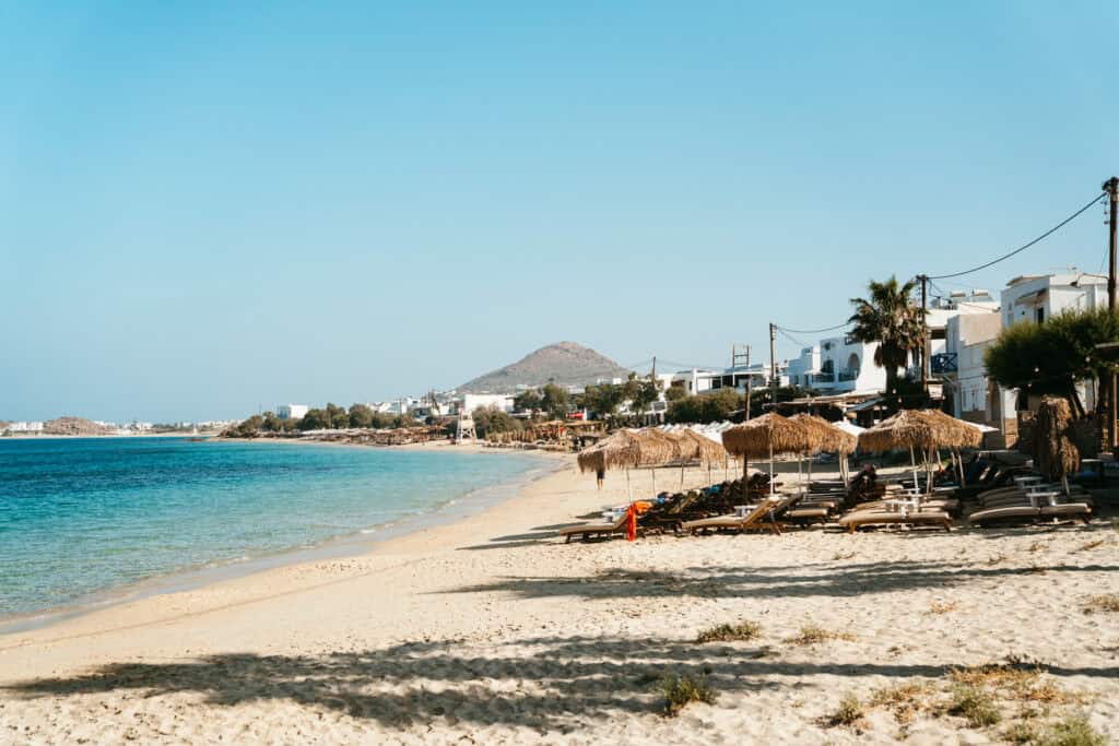 Early morning view of Agia Anna Beach in Naxos with empty sun loungers and thatched umbrellas lining the quiet shoreline, white buildings in the background, and a hill rising in the distance.