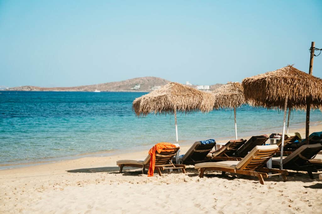 Thatched umbrellas and wooden loungers facing the Aegean Sea on Agia Anna Beach in Naxos, with a quiet shoreline and hilly island landscape in the distance.