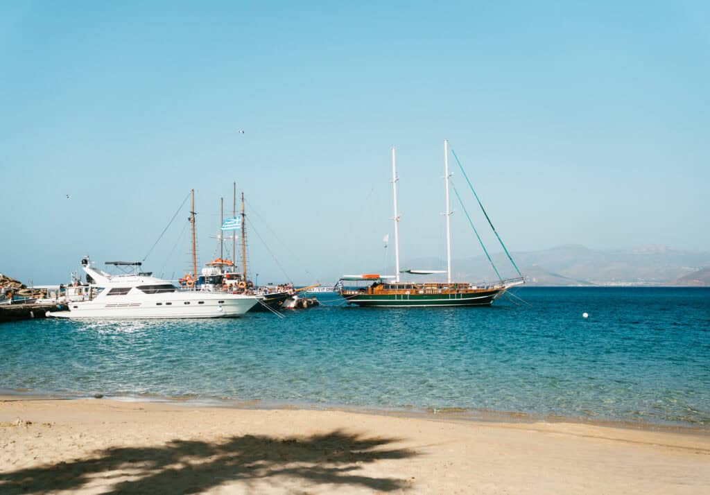 Sailing yachts and tour boats anchored near the shore at Agia Anna, Naxos, with calm turquoise water and hazy mountains in the distance.