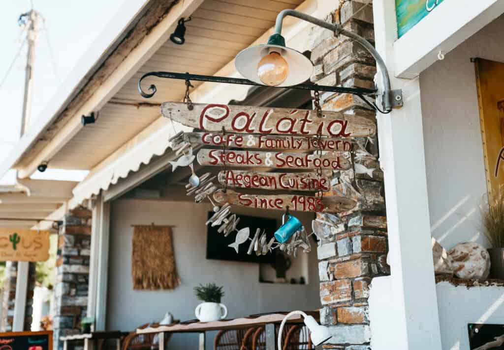 Rustic wooden sign for Palatia Tavern in Agia Anna, Naxos, advertising Aegean cuisine, seafood, and steaks, with fish decorations and a cozy outdoor dining area in the background.