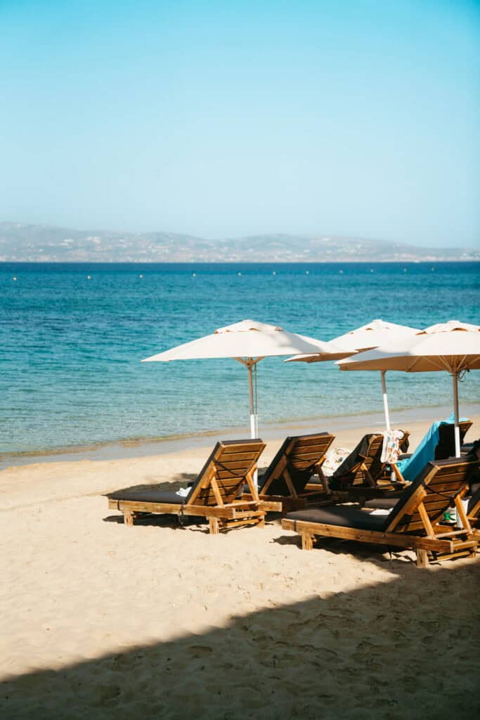 Wooden loungers and white umbrellas on the sandy shore of Agia Anna Beach in Naxos, facing calm turquoise waters and distant hills across the Aegean Sea.