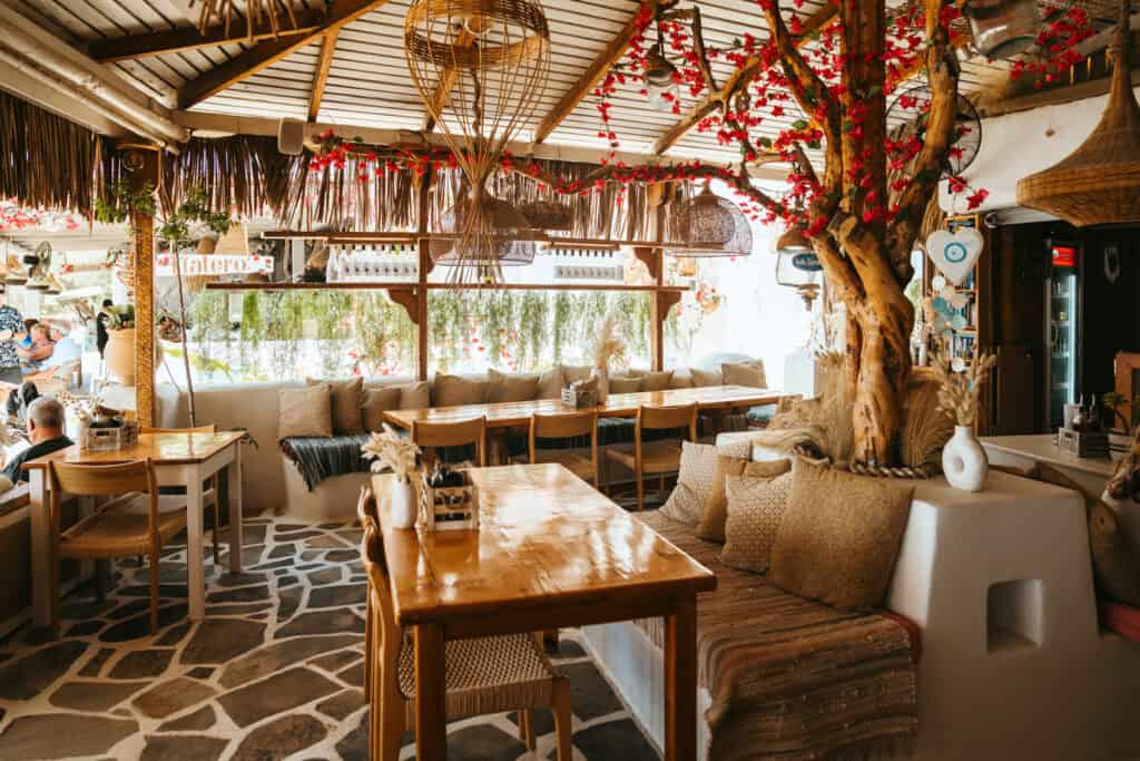 Cozy boho-style restaurant interior in Agia Anna, Naxos, with natural wood tables, woven decor, and a tree wrapped in red flowers as a centerpiece.
