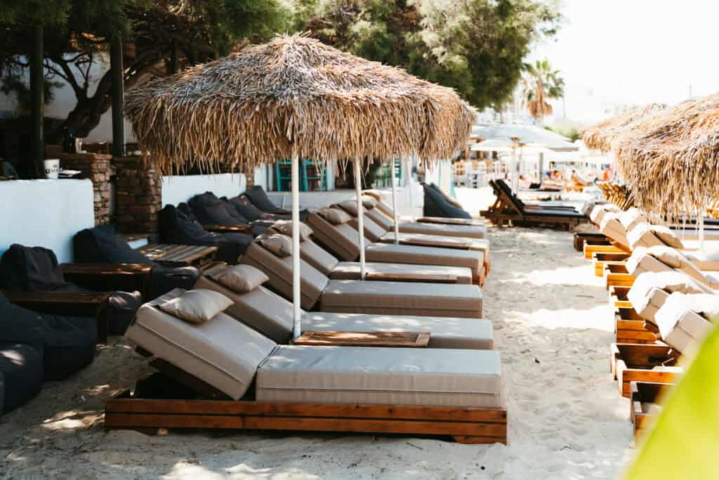 Cushioned sun loungers under thatched umbrellas on the sandy beachfront of Agia Anna, Naxos, with bean bag chairs and a shaded café in the background.