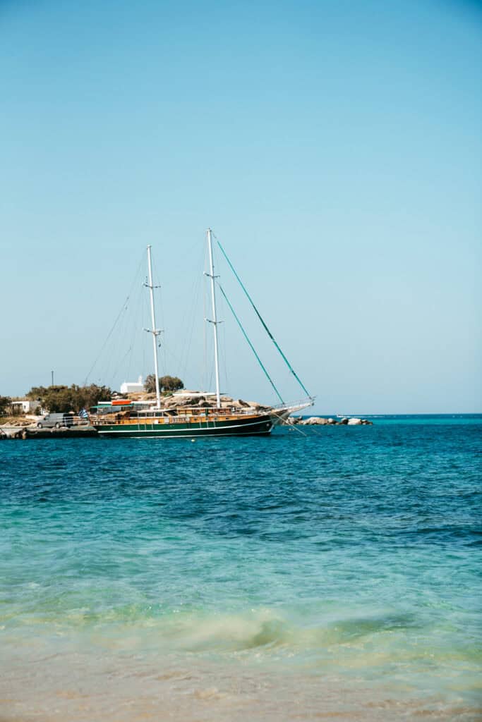 Large wooden sailboat anchored near the shore in Agia Anna, Naxos, with calm turquoise waves in the foreground and a small white chapel on a rocky outcrop in the background.