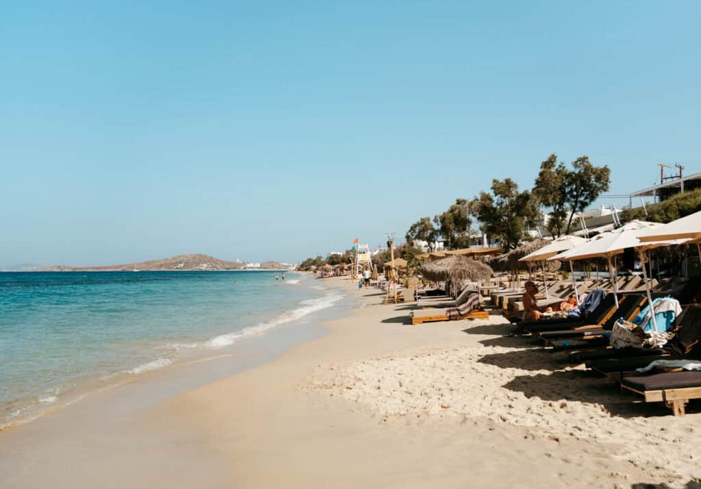 Long stretch of Agia Anna Beach in Naxos with rows of sun loungers and umbrellas lining the sand, gentle waves lapping the shore, and a lifeguard tower in the distance.