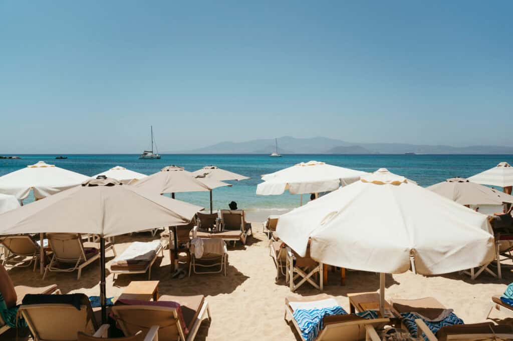 Sunbeds and umbrellas lined up on the sandy shore of Agia Anna Beach in Naxos, with turquoise water, sailboats in the distance, and hazy mountains on the horizon.