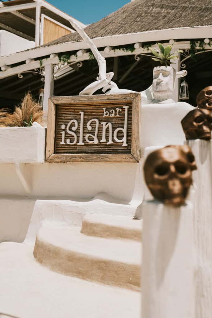 Entrance sign for Island Bar in Agia Anna, Naxos, featuring rustic wooden details, whitewashed steps, and quirky island-themed decor.