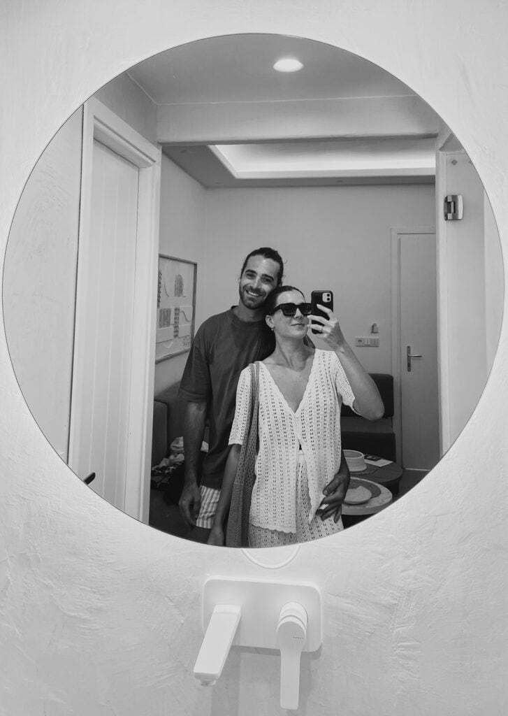 Caleb and Sarah taking a mirror selfie inside a stylish hotel in Agia Anna, Naxos, with minimalist decor, soft lighting, and modern finishes.