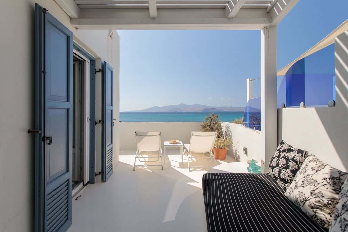 Private balcony at a Iria Beach Art Hotel (seaside) with two lounge chairs and a table facing the Aegean Sea. The white terrace is framed by blue shutters and a black striped daybed with decorative pillows.