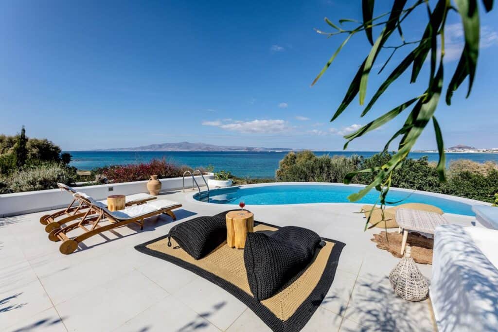 A luxurious seaside patio features a round swimming pool overlooking a serene blue ocean with mountains in the distance. The space includes wooden loungers, black woven bean bag chairs on a tan rug, and a small wooden table with a drink, framed by lush greenery and a clear sky.